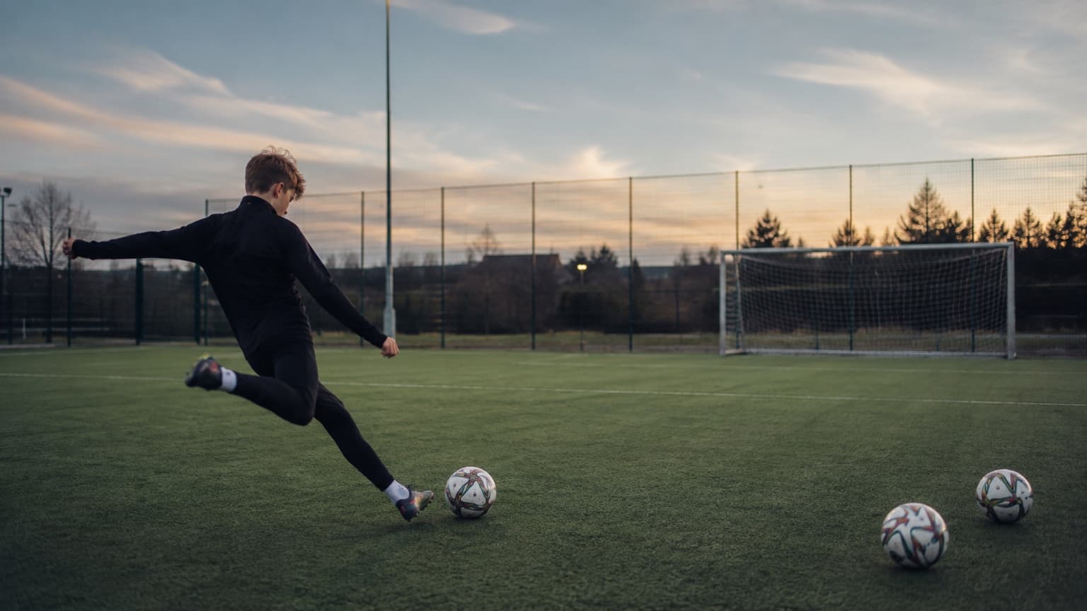 A goal on an artificial pitch at sunset with footballs on the turf in front of it.