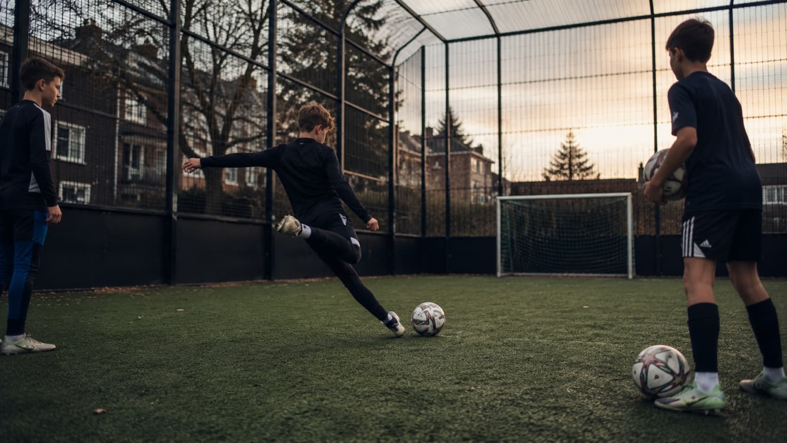 Three young players in a fenced football cage at dusk, one striking the ball towards a small goal.