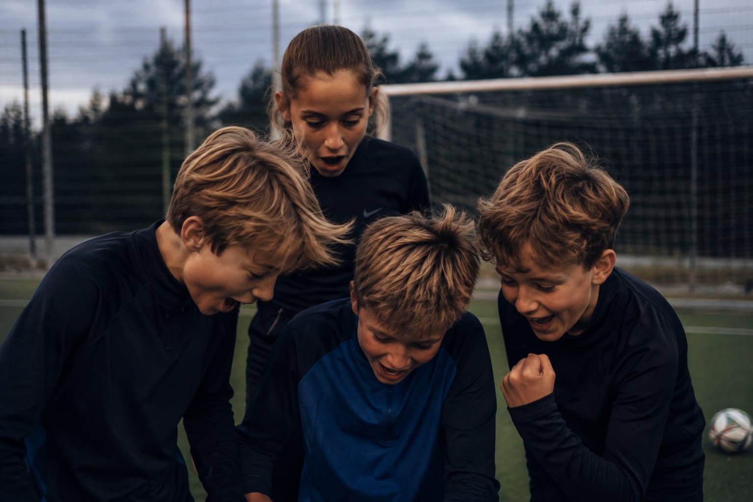Four young players lean in together on a pitch, reacting to a shot-speed result with open mouths and a clenched fist.