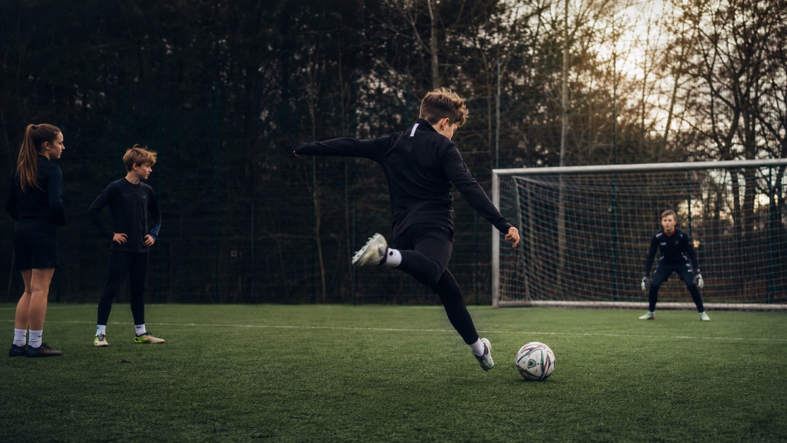 A young player strikes a football towards a goal on an artificial pitch at dusk while a goalkeeper waits and two friends watch.